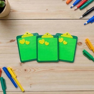 Può includere: Tre lavagne verdi con dettagli a cuore giallo sono disposte su una superficie di legno chiaro. Ogni lavagna ha una clip e un bordo gialli. Penne colorate sono sparse intorno alle lavagne.