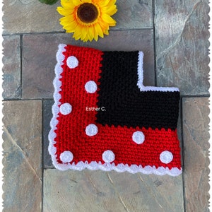 May include: A red and black crocheted baby blanket with white polka dots and a white crocheted border. The blanket is on a stone surface with a sunflower in the background. The text "Esther C." is visible on the blanket.