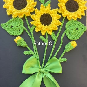 May include: A crocheted sunflower bouquet with green leaves and a green satin bow. The bouquet is on a black background and has the text "Esther C." written on it.