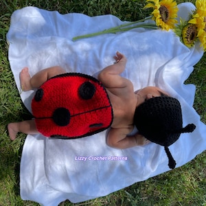 May include: A baby wearing a red and black crocheted ladybug costume with a matching hat. The baby is lying on a white blanket on a bed of green grass. The text "Lizzy Crochet Patterns" is visible in the bottom center of the image.