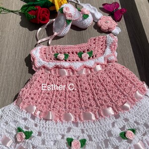 以下が含まれることがあります： ピンクと白のクロシェ編みのベビー ドレス。白いクロシェ編みのヘッドバンド、白いクロシェ編みの靴、そして中央にパールが付いたピンクのクロシェ編みの花が飾られています。ドレスにはピンクのサテンリボンがあしらわれ、白いクロシェ編みのバラと緑の葉が飾られています。