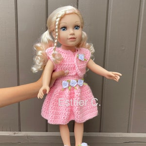 Könnte beinhalten: Ein rosa Häkelkleid mit weißen Blumenapplikationen und passenden Schuhen. Die Puppe hat blondes Haar, das zu Zöpfen mit einem Zopf geflochten ist.