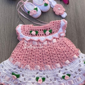 以下が含まれることがあります： ピンクと白のクロシェ編みのベビー ドレス、白いクロシェ編みのヘッドバンド、白いクロシェ編みの靴、ピンクのクロシェ編みの花。ドレスはウエストにピンクのリボン、白いクロシェ編みの裾があります。