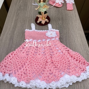 May include: A pink crocheted baby dress with a white crocheted trim and a white flower on the bodice. The dress is paired with a matching pink crocheted headband and a pair of pink crocheted booties with white flowers.