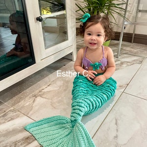 May include: A young child wearing a purple and green mermaid costume with a crocheted tail. The child is sitting on a tiled floor with a white cabinet in the background. The text "Esther C." is written on the floor.