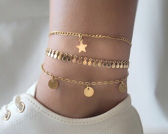 May include: Gold ankle bracelet set with three chains. The top chain has a star charm, the middle chain has small round charms, and the bottom chain has larger round charms.