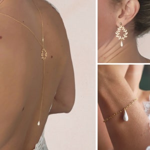 Peut inclure: Un collier en chaîne dorée avec un délicat motif de feuilles et un pendentif en forme de larme blanche. Le collier est porté comme un collier de dos et a un deuxième pendentif en forme de larme en bas. L'image montre également une paire de boucles d'oreilles dorées avec un motif de feuilles et un pendentif en forme de larme blanche, ainsi qu'un bracelet doré avec un pendentif en forme de larme blanche.