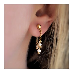 Op de afbeelding: Gouden oorhangers met een kleine gouden bol aan de bovenkant en een ketting die naar een cluster van kleine witte parels leidt.
