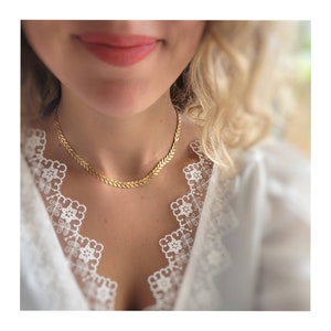 Könnte beinhalten: Eine goldene Kettenkette mit einem Chevron-Muster. Die Kette wird von einer Person mit blonden Haaren getragen und sitzt vor einem weißen Spitzenhintergrund.