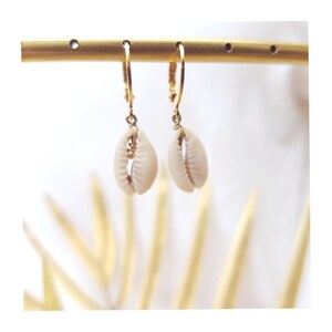 Puede incluir: Aretes de aro dorados con un dije de concha de cauri blanca colgando de cada aro.