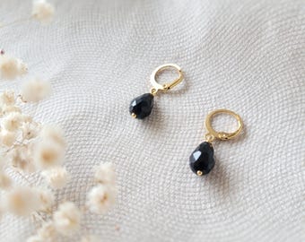 May include: A pair of gold-colored hoop earrings, each with a teardrop-shaped black gemstone pendant. The earrings are displayed on a white, textured surface, with soft, blurred floral accents in the background.