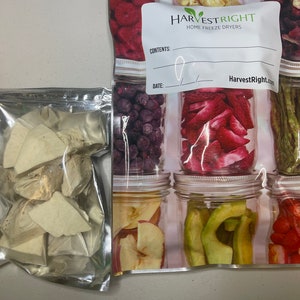 May include: A clear plastic bag with a label that reads "HarvestRight Home Freeze Dryers" and a handwritten date. The bag contains dried fruit slices in various colors, including red, green, and purple. The bag is sitting on a white surface.