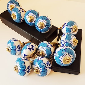 May include: A set of 12 blue and white ceramic cabinet knobs with floral designs. The knobs have a gold-coloured metal base and are arranged on a black surface.