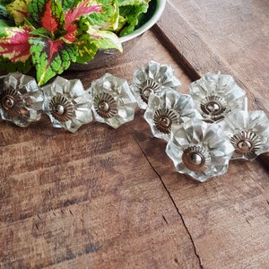 Peut inclure: Un ensemble de huit boutons de cabinet en verre transparent avec une finition argentée. Les boutons ont une forme de fleur avec un centre surélevé.