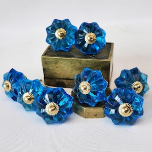 Op de afbeelding: Een verzameling van acht blauwe, bloemvormige kastknoppen met gouden centra. De facetknoppen worden getoond op een messingkleurige doos en een kleine messingkleurige standaard. De knoppen staan tegen een witte achtergrond. Diameter ca. 4 cm.