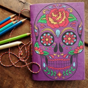 May include: Purple notebook with a colorful sugar skull design. The skull has a flower on top and is surrounded by floral details.