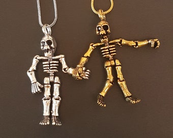 May include: Two skeleton pendant necklaces, one silver and one gold, are shown hanging from their chains. The skeletons are holding hands.