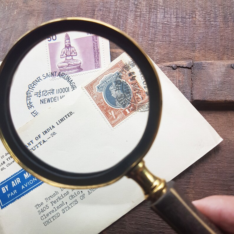 May include: A magnifying glass is positioned over a vintage envelope with three stamps. The stamps are from India and feature a person's portrait and a seated figure. The envelope has a blue and white label that reads "By Air Mail Par Avion".