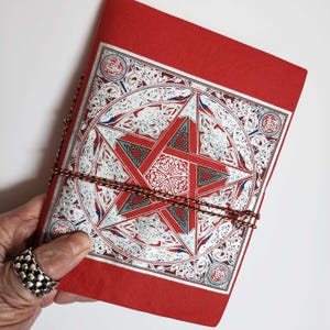Könnte beinhalten: Ein rotes Notizbuch mit einem dekorativen Pentagramm-Design auf dem Einband. Der Einband weist ein detailliertes silbernes, rotes und blaues Muster auf. Das Notizbuch ist mit einer braunen und schwarzen Kordel gebunden.