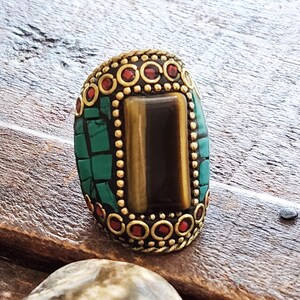 May include: An oval-shaped ring with a central rectangular tiger's eye stone. The ring is framed by a gold-colored metal setting with red and green accents. The ring's design features a mosaic-like pattern with small, square green stones.