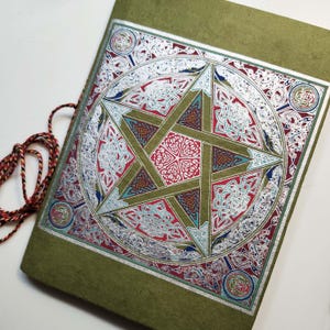 Puede incluir: Un diario verde con un diseño de pentagrama plateado. El pentagrama está rodeado de intrincados nudos plateados y detalles en rojo, azul y verde. Una cuerda multicolor está unida al diario.