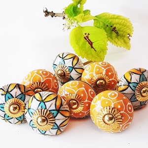 Może przedstawiać: Zestaw ośmiu ceramicznych gałek z kwiatowym wzorem w kolorze żółtym, białym i niebieskim. Gałki mają złoty metalowy akcent w środku.