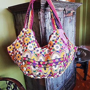 May include: A hobo-style shoulder bag featuring vibrant, multi-coloured embroidery in shades of yellow, pink, blue, and green. The fabric is adorned with small circular mirrors and white cowrie shells, with a wide, matching embroidered shoulder strap.