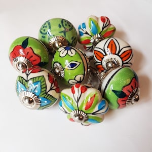 May include: A set of eight colorful ceramic cabinet knobs with floral designs. The knobs are green, white, red, orange, and blue. Each knob has a silver metal base.