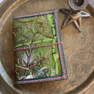 May include: A green journal with detailed embroidery and sequins. The cover displays a floral design with gold and silver accents. The journal is bound with a decorative cord and has a colourful border.