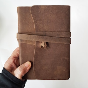 May include: A brown leather journal with a wrap-around strap closure. The journal is rectangular and appears to be handmade, with a rustic, textured appearance. The strap is secured with a small leather loop.