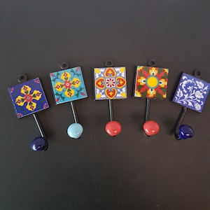 Puede incluir: Cinco ganchos decorativos con azulejos de cerámica coloridos que presentan patrones geométricos. Los azulejos son en tonos de azul, rojo, amarillo y blanco. Los ganchos son de metal negro con un pomo redondo en la parte inferior.