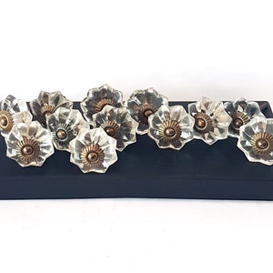 Peut inclure: Un ensemble de dix boutons de cabinet en verre transparent en forme de fleur avec des accents en laiton. Les boutons sont disposés sur une surface noire.
