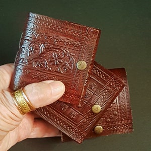 Op de afbeelding: Een set van drie bruine leren onderzetters met reliëf bloemen- en geometrische patronen. Elke onderzetter heeft een messing knoopsluiting. De onderzetters worden in een hand gehouden, waarbij het gedetailleerde ontwerp wordt benadrukt.