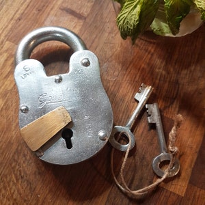May include: A silver padlock with a brass keyhole and a brass plate with the text "Good Luck" on it. The padlock is attached to two keys with a brown string.