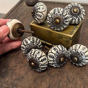 Puede incluir: Una colección de pomos redondos de cerámica para armarios con base color crema, intrincados patrones florales negros y puntos blancos en relieve. Cada pomo tiene un centro metálico oscuro y un perno roscado para su instalación.