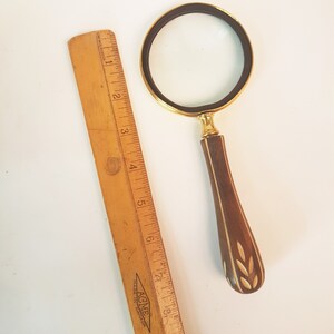 May include: A magnifying glass with a gold-tone frame and a wooden handle with a carved design. The handle has a white inlay with a leaf pattern. The magnifying glass is on a white surface.