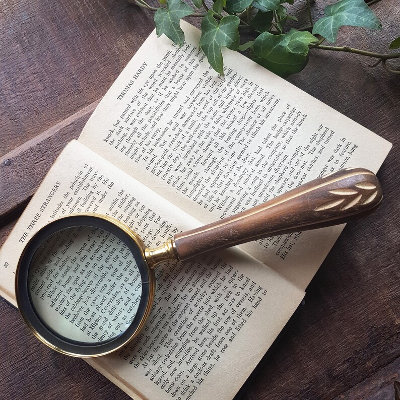 May include: A magnifying glass with a gold-tone frame and a wooden handle with a carved design sits on an open book. The book is titled "The Three Strangers" and features text by Thomas Hardy.