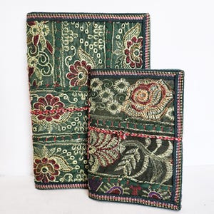 Könnte beinhalten: Zwei dekorative Journale mit gestickten Blumenmustern. Die Journale haben einen dunkelgrünen Hintergrund mit roten, goldenen und cremefarbenen Stickereien. Die Ränder sind mit einer dunkelgrünen und roten Bordüre versehen. Die Journale sind mit einer rot-cremefarbenen Schnur gebunden.