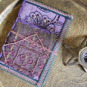 Può includere: Un diario fatto a mano con un design floreale viola sulla copertina. Il diario è adornato da ricami intricati, perline e un bordo decorativo. Un cordino colorato avvolge il diario per tenerlo chiuso. Il diario è su una superficie color oro.