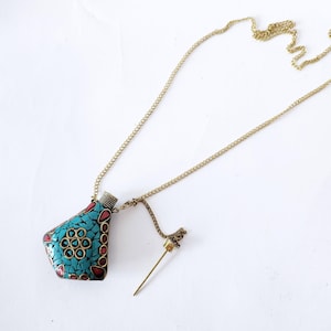 Könnte beinhalten: Eine goldene Kettenkette mit einem türkisfarbenen und roten Mosaik-Anhänger in Form einer Flasche. Der Anhänger hat eine kleine Öffnung oben und eine goldene Kette ist daran befestigt. Die Kette ist lang genug, um als Halskette getragen zu werden. Der Anhänger ist auch an einer separaten Kette mit einer kleinen goldenen Nadel befestigt.