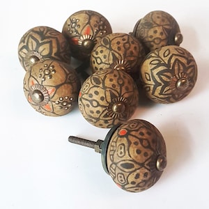 Op de afbeelding: Acht bruine keramische kastknoppen met ingewikkelde zwarte en oranje bloemmotieven. Elke knop heeft een schroef van messing en een messing accent in het midden.