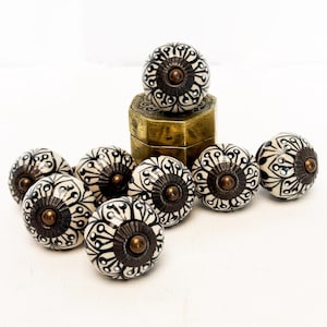 Peut inclure: Un ensemble de huit boutons de placard ronds en céramique avec un motif floral noir et blanc et une pièce centrale en métal couleur bronze. Un bouton est placé sur une petite boîte hexagonale en métal couleur laiton vieilli.