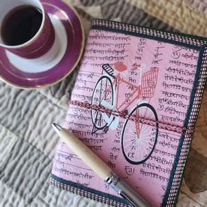 Könnte beinhalten: Ein rosa Notizbuch mit einer Fahrradillustration und Schrift, gebunden mit rot-weißem Garn, liegt neben einem Stift und einer Tasse Kaffee. Das Cover des Journals zeigt ein Fahrraddesign und einen dekorativen Rand.