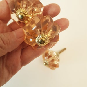 May include: Three peach-coloured glass cabinet knobs with gold accents. The knobs have a fluted design and are held in a hand.