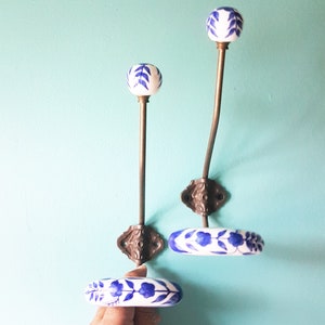 Peut inclure: Deux patères murales décoratives avec des boutons en céramique florale blanche et bleue. Les crochets ont une finition bronze et sont montés sur un mur bleu clair. Un crochet est droit, l'autre est incurvé. Une main tient une poignée en céramique assortie.