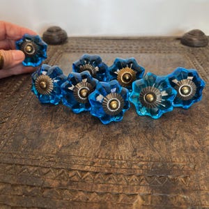 May include: A collection of eight faceted blue glass cabinet knobs, each featuring a star-shaped design with a central metallic brass-toned circular base and a small protruding screw head in the middle, arranged in a cluster on a flat surface.