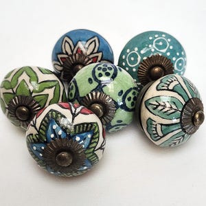 May include: A selection of seven ceramic cabinet knobs. The knobs are decorated with floral and geometric patterns in blue, green, and white. Each knob has a bronze-coloured metal base.