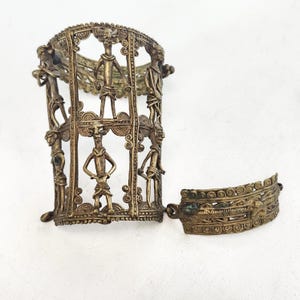 May include: Two-piece antique brass bracelet set. The larger piece is a cage-like structure with stylized human figures. The smaller piece is a curved band with decorative patterns. Both pieces have a tarnished, aged appearance.