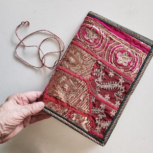 Op de afbeelding: Een rechthoekig stoffen tasje met een patchworkontwerp van magenta en donkerrode panelen. Het oppervlak is rijkelijk versierd met goudkleurige pailletten en ingewikkeld metaaldraadborduurwerk. Een dun, gedraaid koord is aan de bovenrand bevestigd.