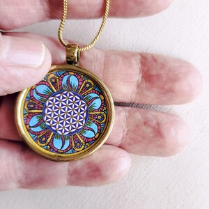 Peut inclure: Un pendentif rond doré avec un motif géométrique central de fleur de vie en blanc sur fond violet, entouré d'une bordure décorative de formes en forme de pétales bleues, jaunes et rouges, le tout enchâssé dans un cadre en métal doré.